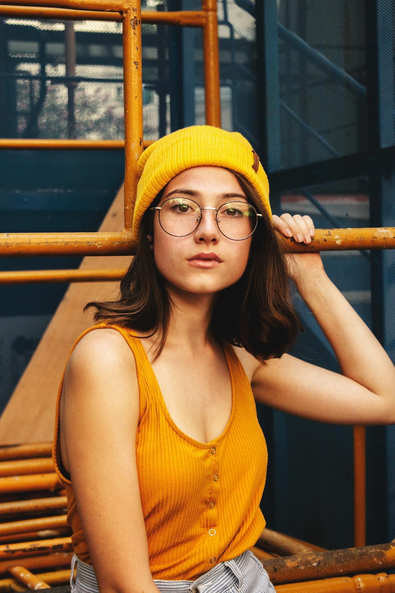 Stylish woman posing in mustard beanie and glasses against industrial backdrop.
